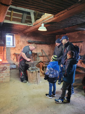 2 Männer und 2 Jungs sehen dem Schmied bei der Arbeit im Agroneum zu.