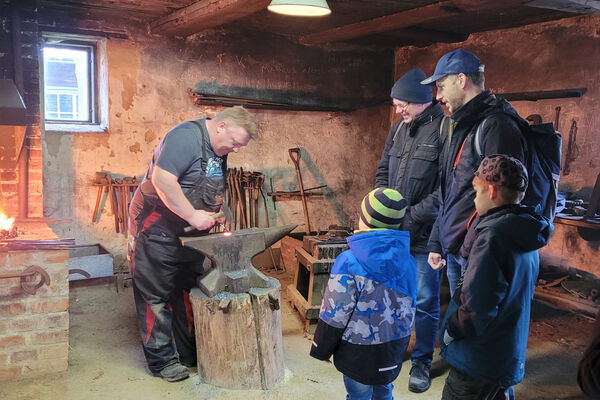 2 Männer und 2 Jungs sehen dem Schmied bei der Arbeit im Agroneum zu.