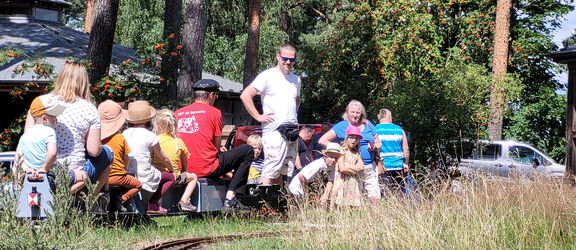 Lokführer und mehrere Kinder sitzen auf einer Modelleisenbahn im Agroneum.