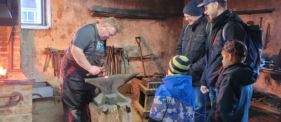 2 Männer und 2 Jungs sehen dem Schmied bei der Arbeit im Agroneum zu.