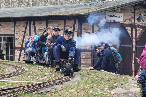 Lokführer und mehrere Kinder sitzen auf einer Modelleisenbahn im Agroneum.