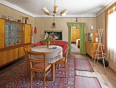 in der Mitte ein Tisch mit 4 Stühlen, links daneben Vitrine, rechts Fenster mit Gardine