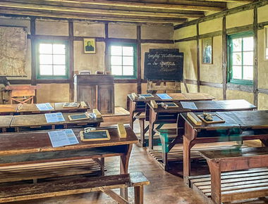 Klassenzimmer vor 150 Jahren, einige Holztische und Bänke mit Holztafeln, Lehrerpodest, Tafel